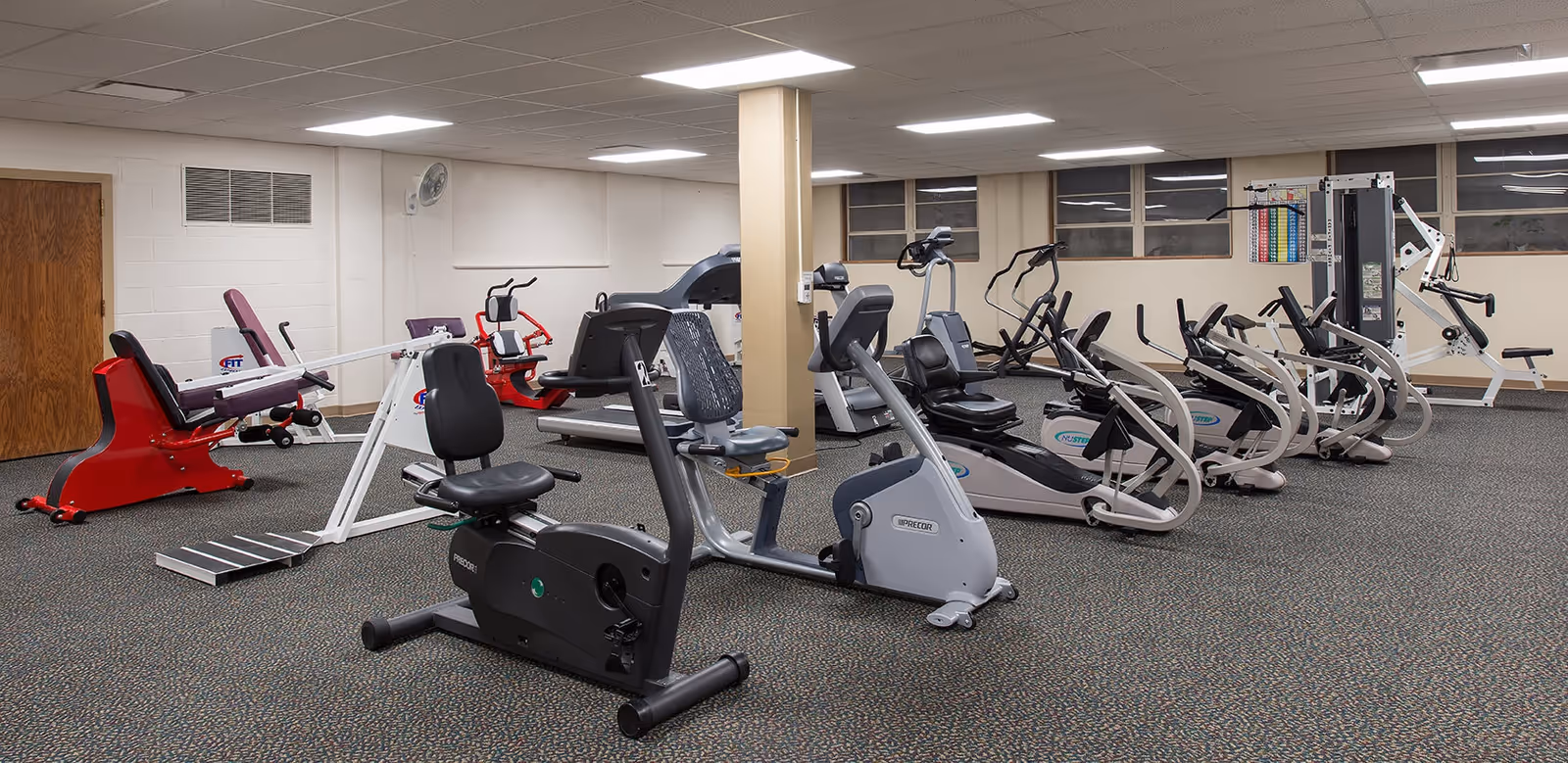 Interior view of a fitness room with various exercise equipment including recumbent bikes, elliptical machines, and strength training machines. The room has carpeted flooring, beige walls, and fluorescent ceiling lights.