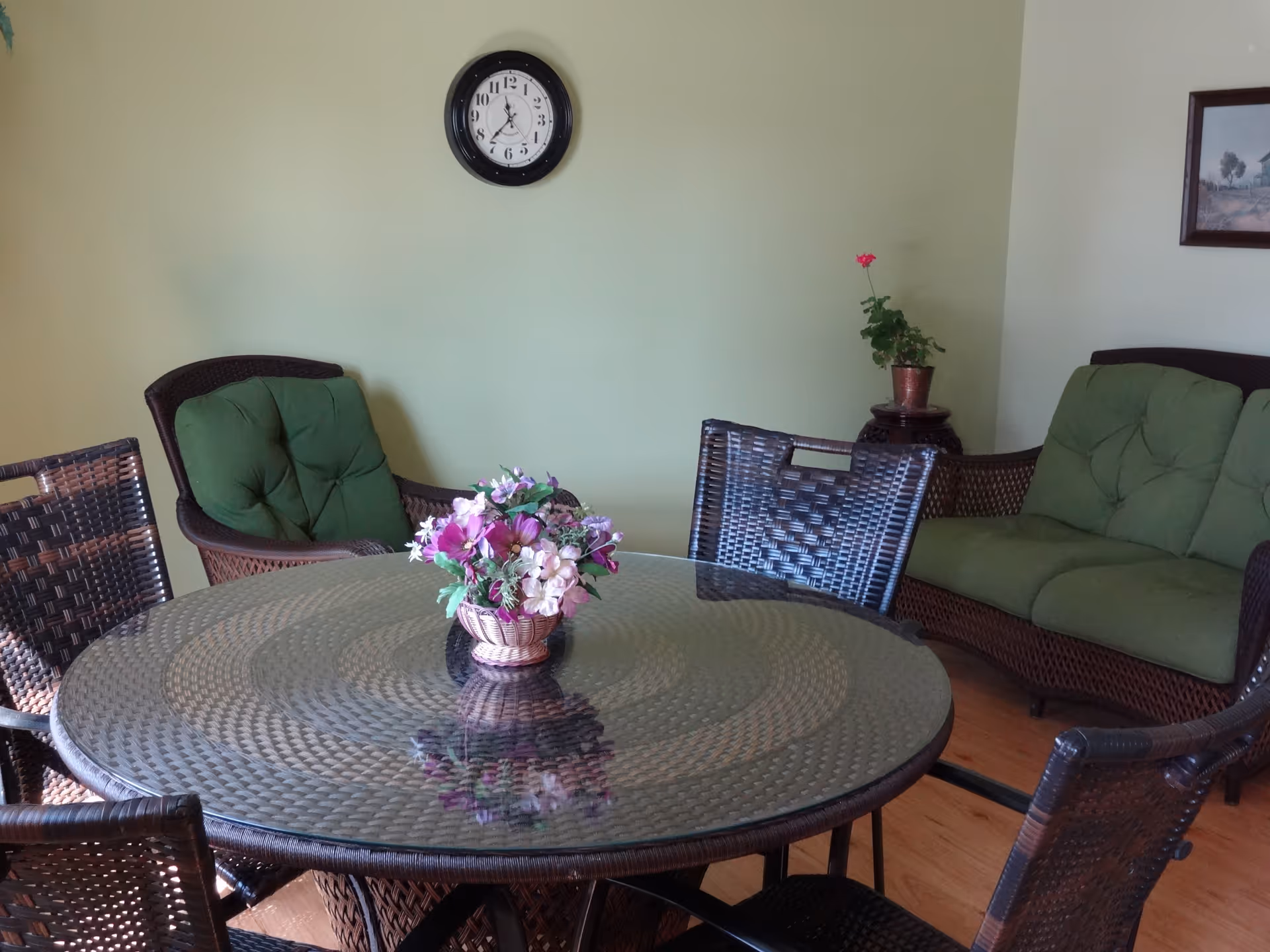 Round glass-top wicker table with a floral centerpiece surrounded by wicker chairs and green-cushioned seating against a light green wall with a clock.