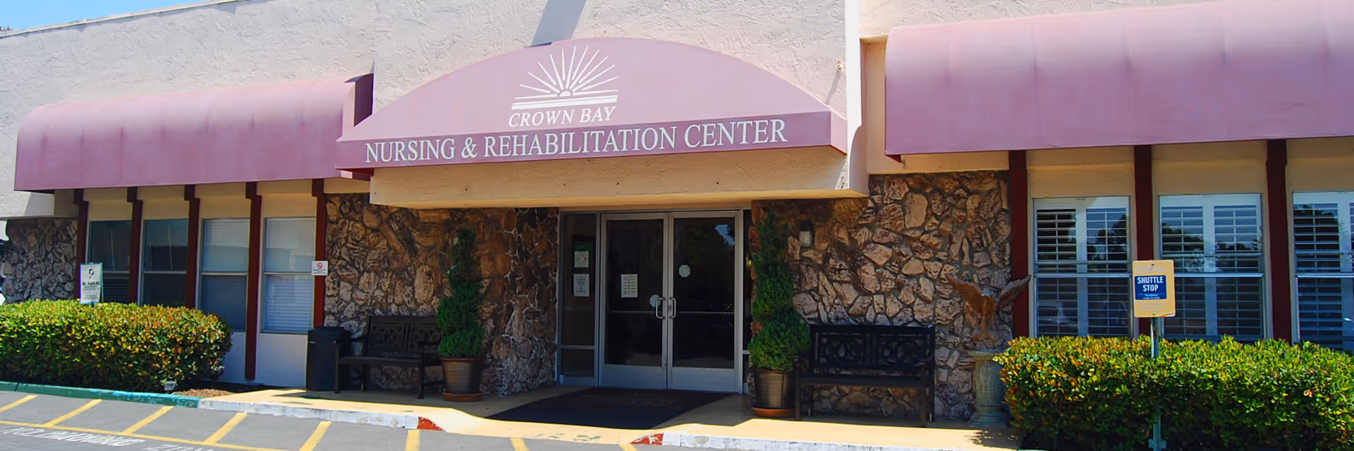 Front entrance of Crown Bay Nursing & Rehabilitation Center featuring a stone facade, double glass doors, two black benches, potted plants, and a purple awning with the facility name. There are bushes on either side and a shuttle stop sign on the right.
