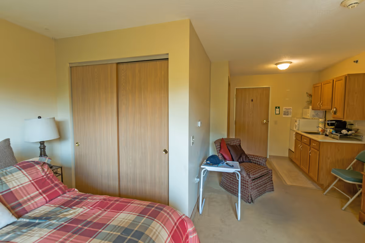 Interior view of a senior living facility room at Heritage Woods of Batavia featuring a bed with a red and beige plaid bedspread, a bedside lamp on a small table, a wooden closet with sliding doors, a patterned armchair with pillows, a small white table holding a hat and remote control, and a kitchenette area with wooden cabinets, a microwave, and a refrigerator.