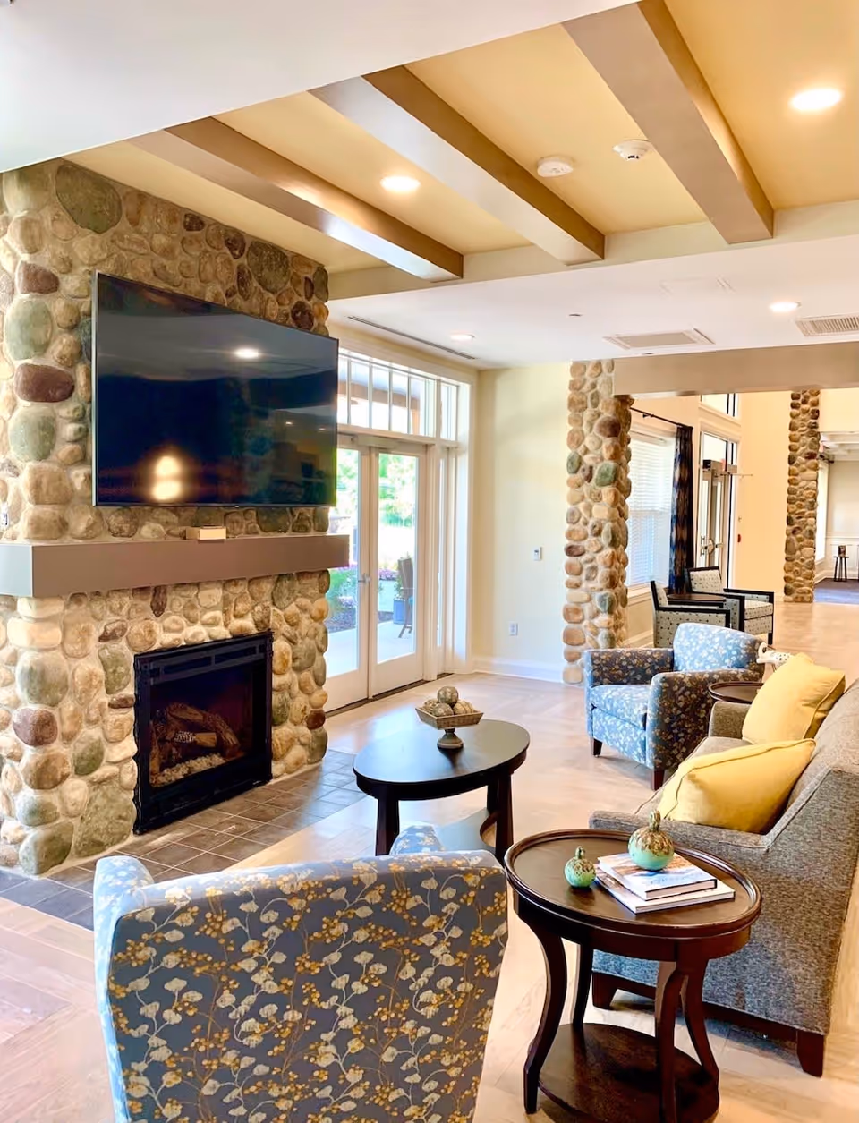 A cozy living room area in a senior living facility featuring a stone fireplace with a mounted flat-screen TV above it. The room has wooden ceiling beams, large windows and glass doors letting in natural light, and comfortable seating including a gray sofa with yellow pillows and two blue floral armchairs. There are two round wooden tables, one with decorative items and books, and the other with a bowl of decorative spheres.
