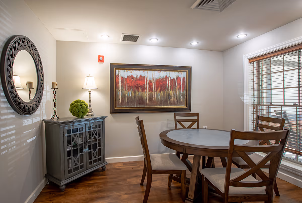 Cozy dining area with a round wooden table and four chairs, a side cabinet with a lamp and decorative items, a round wall mirror, and a framed painting.