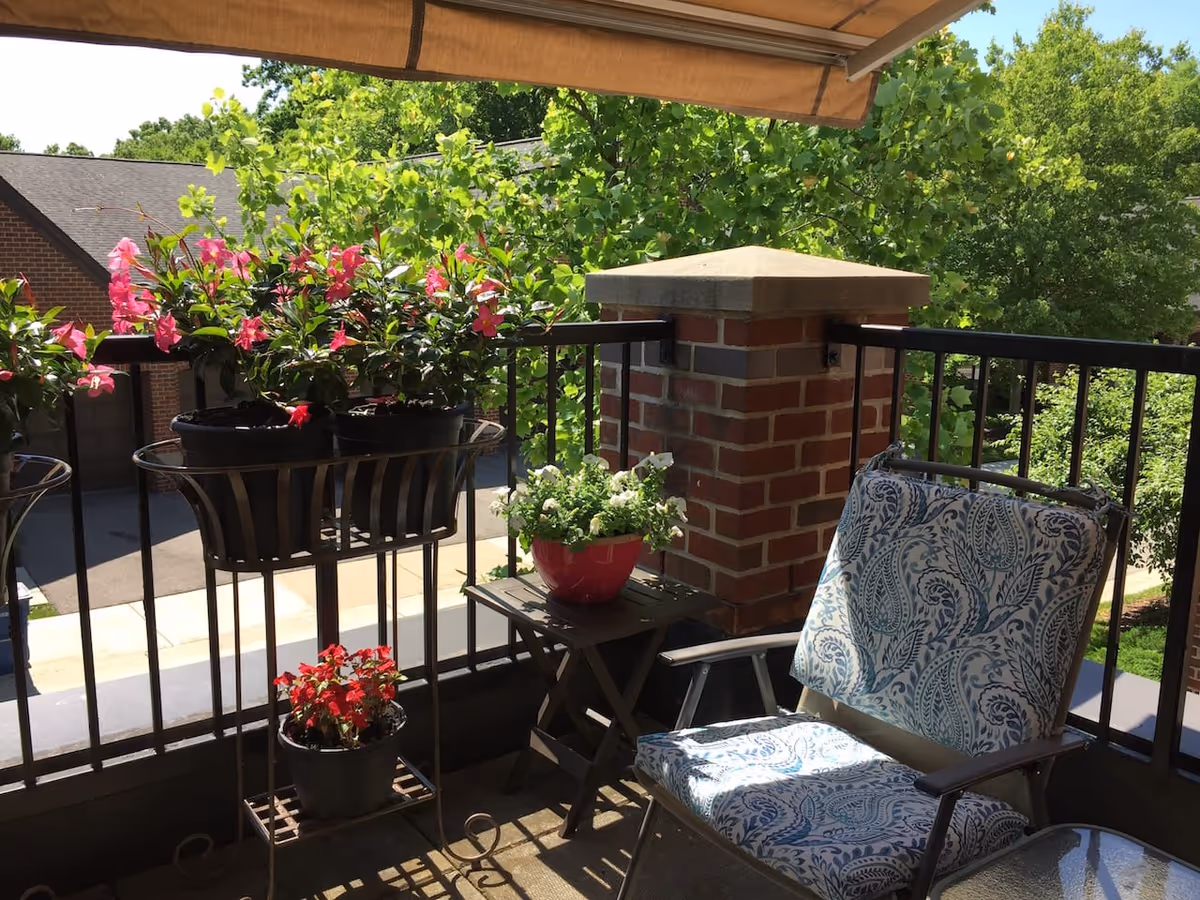 A small outdoor balcony area with a cushioned chair featuring a blue and white paisley pattern, a small table with a red flower pot containing white flowers, and a metal plant stand holding three pots of pink and red flowers. The balcony has a black metal railing and a brick pillar, with green trees and a building visible in the background under a sunny sky.