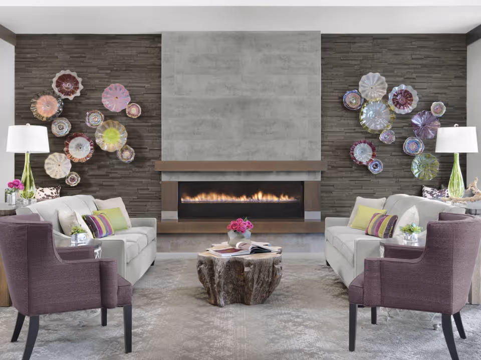 Contemporary lounge with two sofas facing a modern fireplace, purple armchairs, a tree-stump coffee table, and colorful glass wall sculptures.