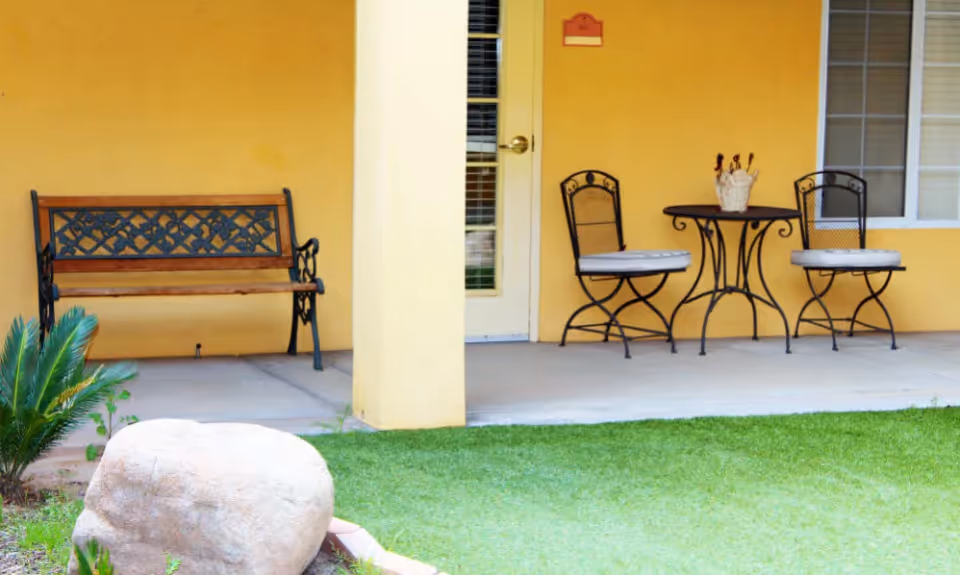 Yellow exterior patio with a bench, a small table and two chairs next to a door overlooking a grassy lawn and decorative rock.