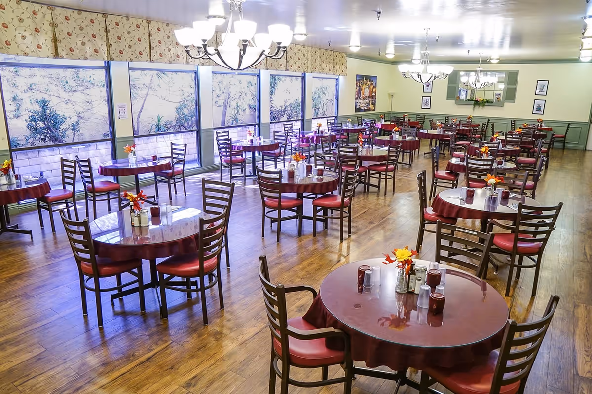 A spacious dining room with multiple round tables covered with maroon tablecloths and surrounded by wooden chairs with red cushions. Each table has a small flower arrangement and condiments. Large windows along one wall let in natural light, and chandeliers hang from the ceiling. The room has wooden flooring and light green walls with framed pictures and a mirror.