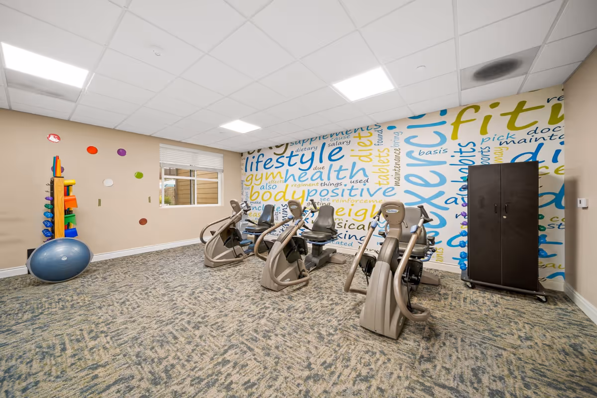 A fitness room with three recumbent exercise bikes positioned on a carpeted floor. The back wall features a colorful mural with words related to health, fitness, and lifestyle. To the left, there is a rack holding various exercise equipment such as dumbbells and mats, and a large blue exercise ball rests on the floor nearby. A window with blinds lets in natural light, and a tall black cabinet stands against the right wall.
