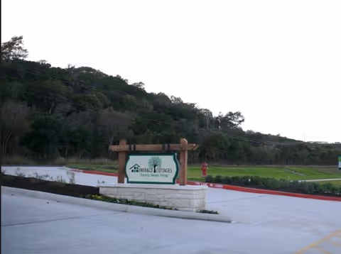 Entrance sign for Emerald Cottages of Kerrville on a stone base in a parking area with a grassy hillside behind it.