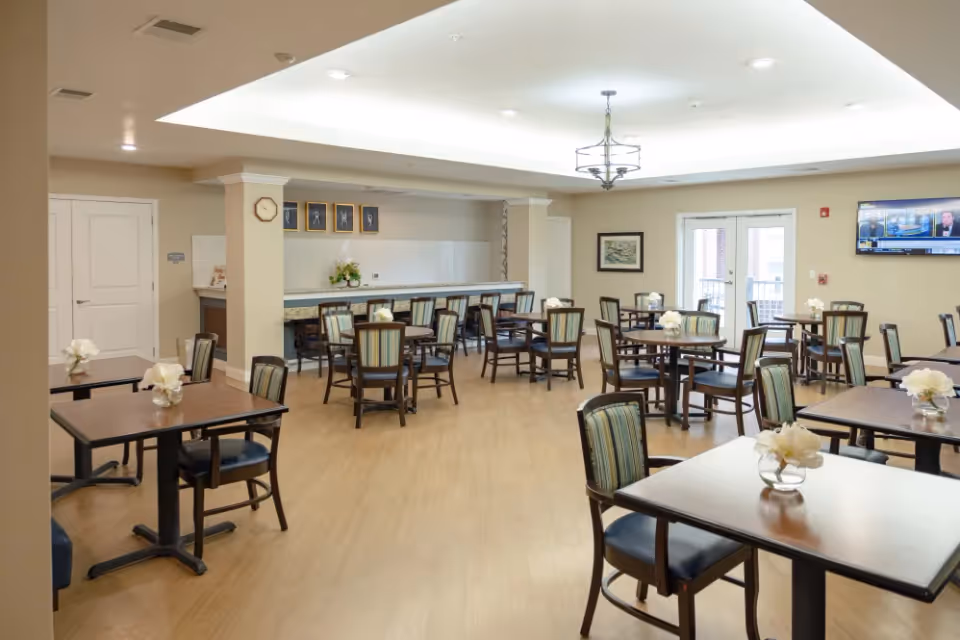Bright senior living dining room with multiple tables and chairs and a service counter at the back.