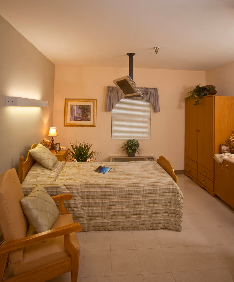 A cozy bedroom in a senior living facility with a single bed covered in a striped bedspread, a wooden armchair with a cushion, a nightstand with a lamp and framed photos, a window with blinds and a valance, a ceiling-mounted TV, and wooden cabinets with decorative plants.