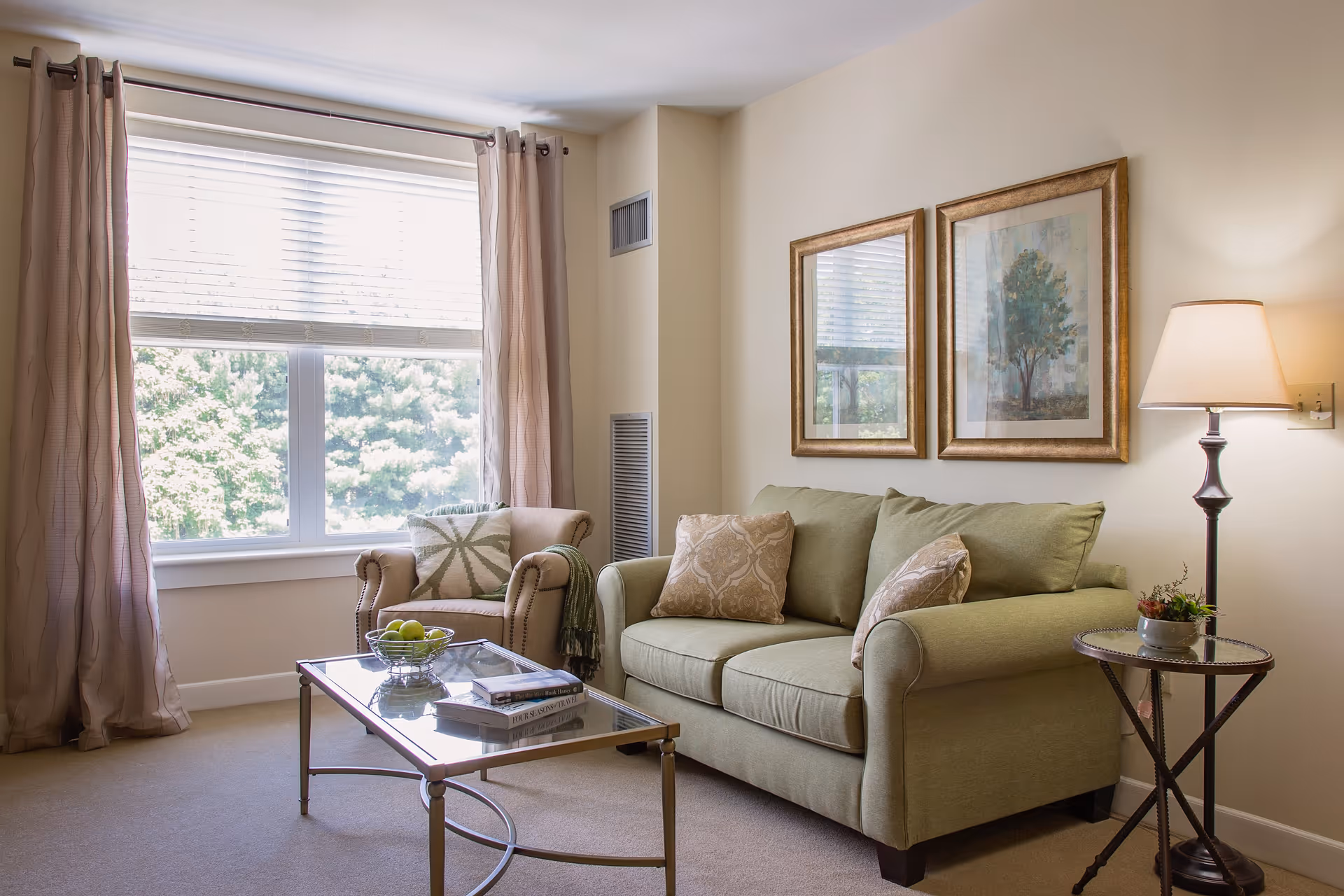 A cozy living room with a green sofa adorned with patterned cushions, a beige armchair with a decorative pillow, a glass coffee table holding a bowl of green apples and books, two framed pictures on the wall, a floor lamp, and large windows with beige curtains letting in natural light.