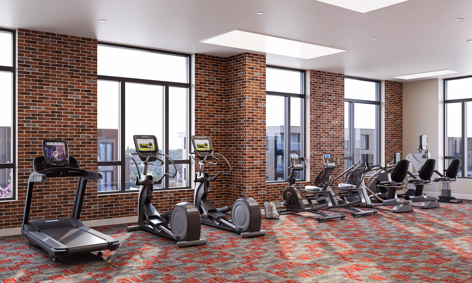 A modern fitness room with large windows and exposed brick walls. The room contains various exercise machines including a treadmill, elliptical trainers, stationary bikes, and recumbent bikes, all arranged in a row on a patterned carpeted floor.