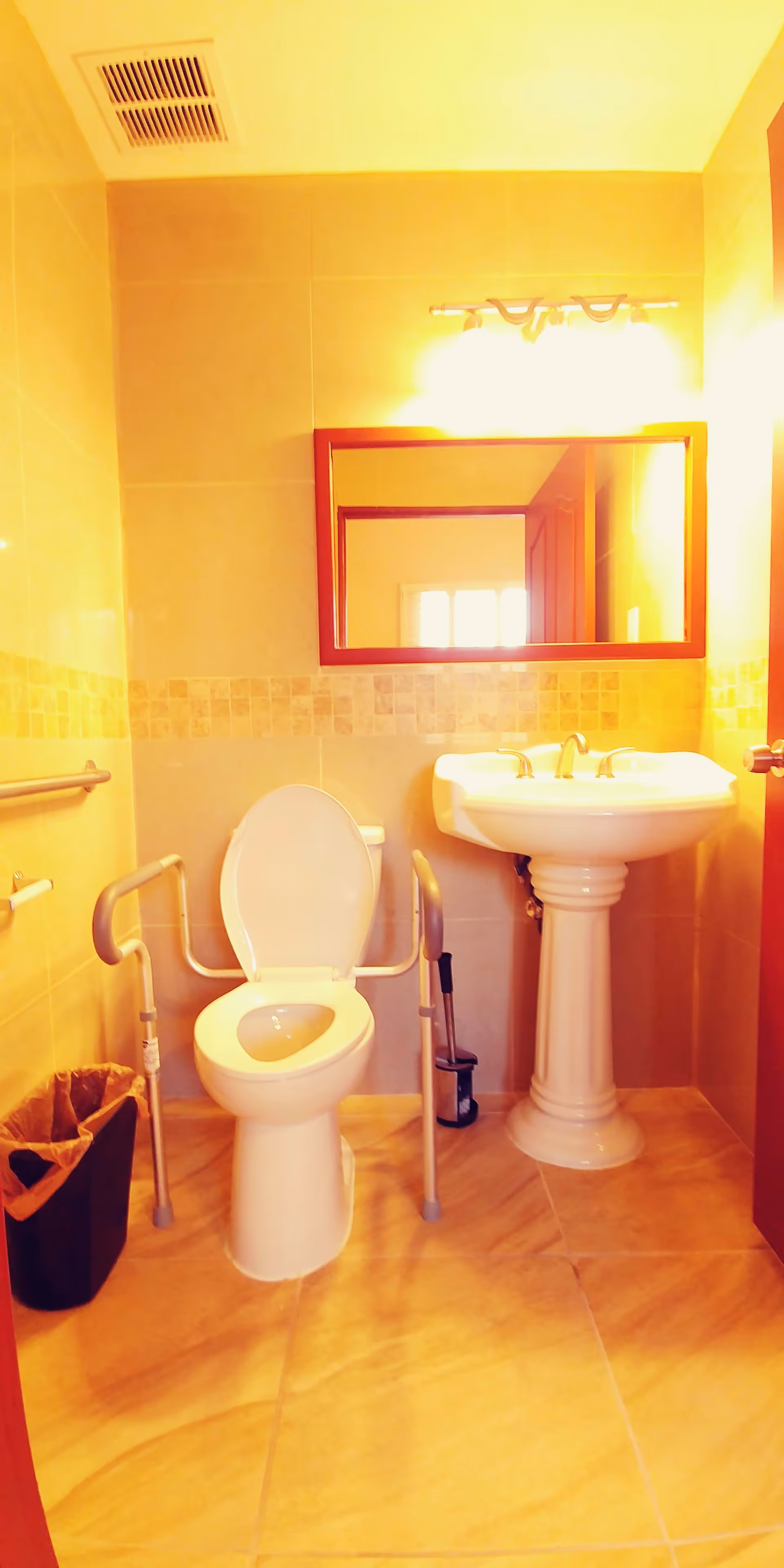 A small bathroom with beige tiled walls and floor. It features a white toilet with safety grab bars on both sides, a white pedestal sink with a mirror above it, and a trash bin to the left of the toilet. There is a light fixture above the mirror and a ventilation vent on the ceiling.
