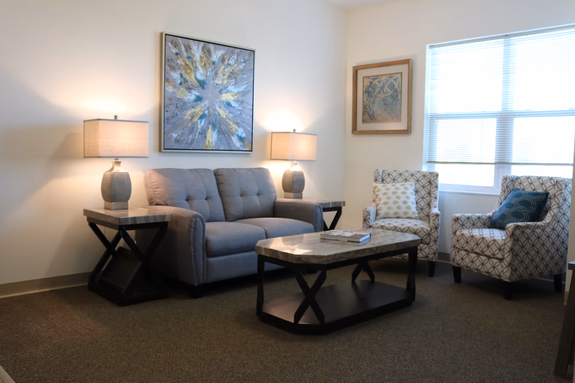 Well-lit sitting area with a gray sofa, two patterned armchairs, a coffee table and side tables with lamps beneath wall art.
