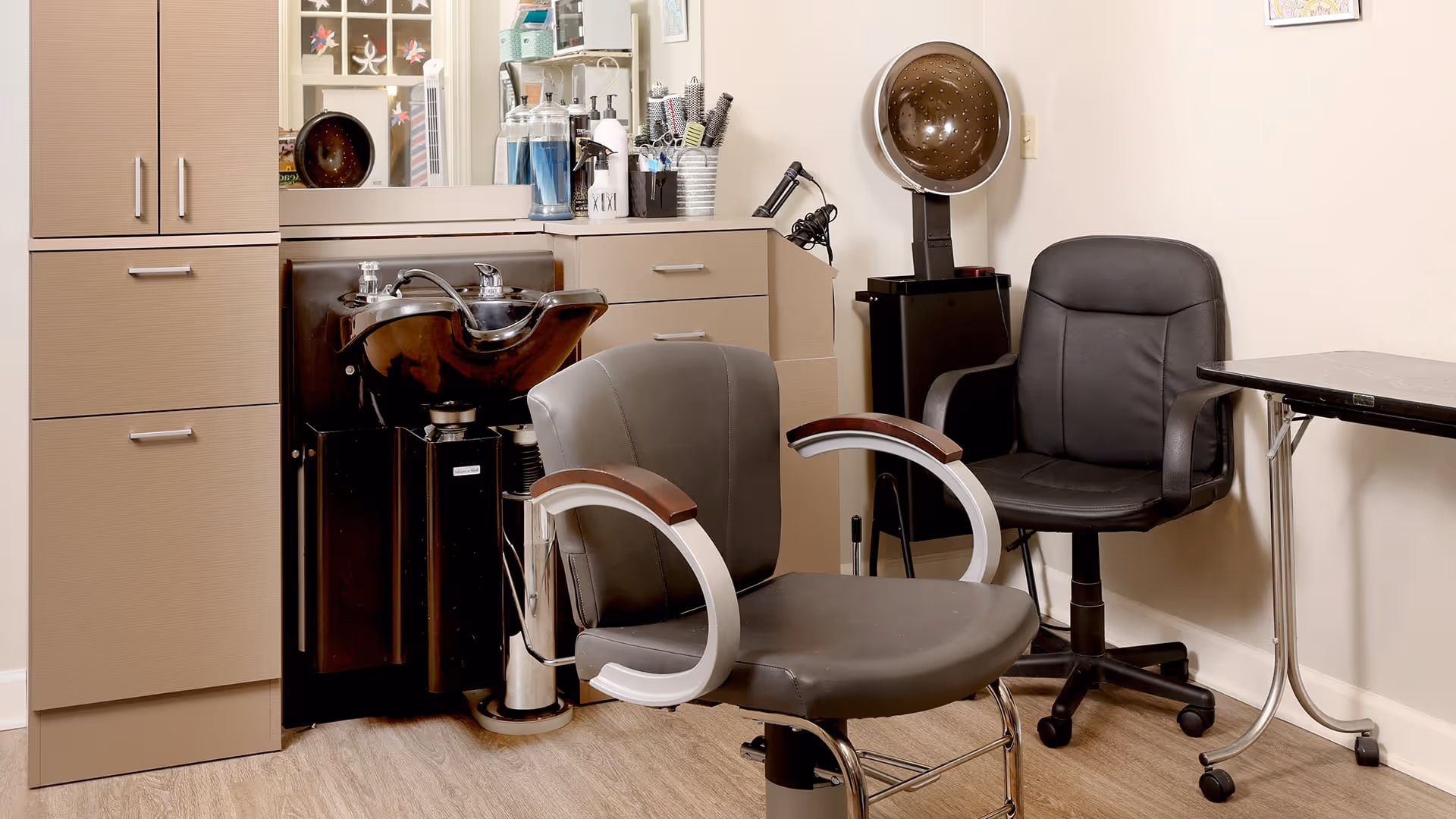 Interior view of a salon area with a black salon chair in front of a hair washing station, a black rolling chair, a hair dryer hood, a small table on wheels, and cabinets with various hair care products on the countertop.