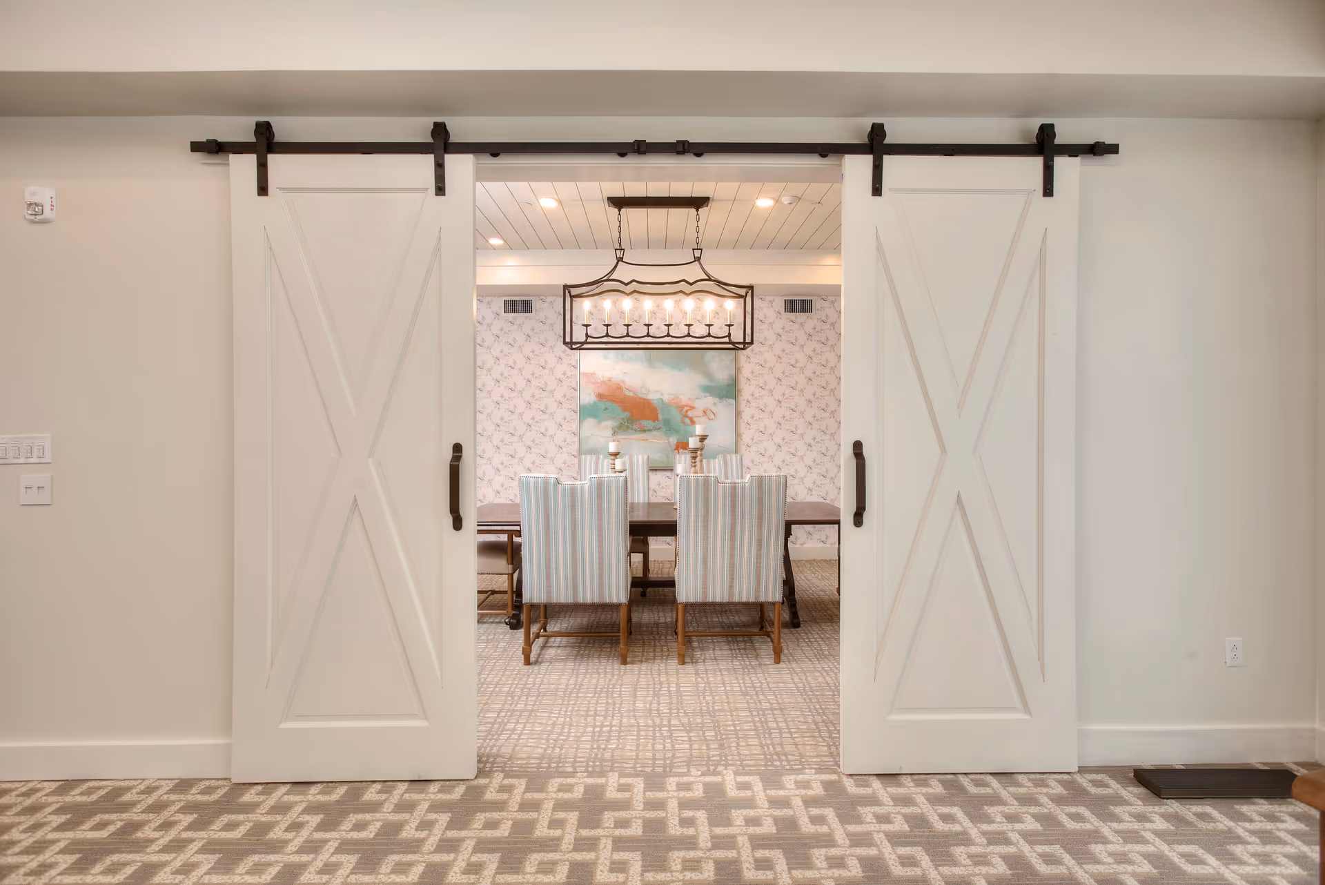 View through two white sliding barn doors into a dining room with a rectangular wooden table surrounded by six striped upholstered chairs. A large rectangular chandelier with multiple lights hangs above the table. The back wall features floral wallpaper and a large abstract painting with teal, white, and rust colors. The floor is carpeted with a geometric pattern.