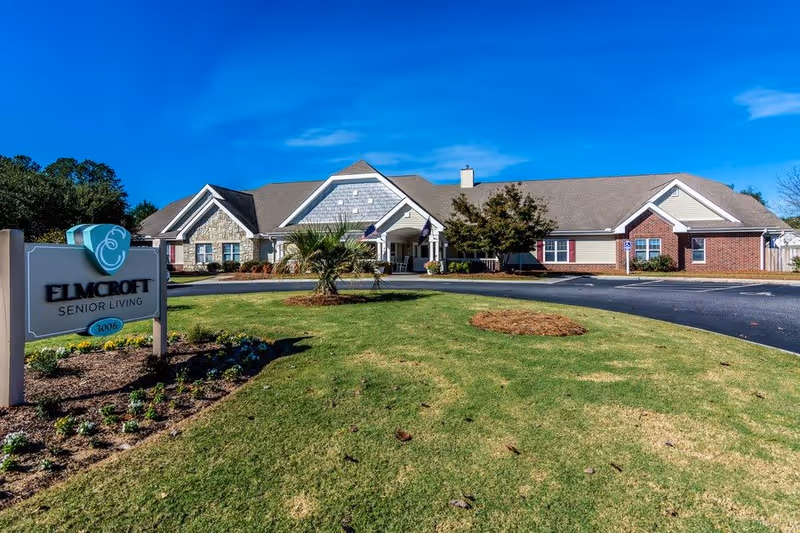 Single-story senior living building with a manicured lawn and an 'Elmcroft Senior Living' sign in the foreground under a bright blue sky.