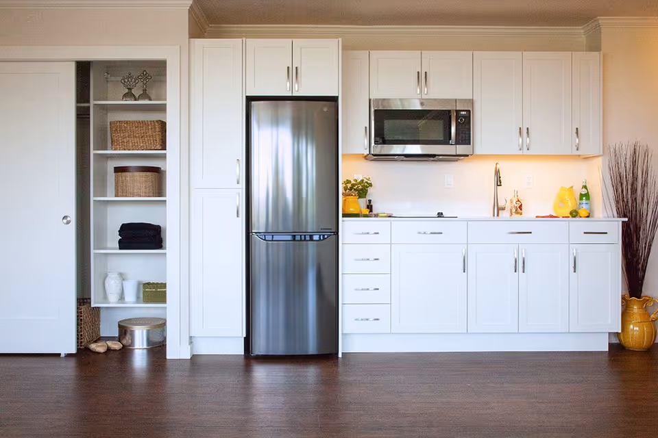 Bright modern kitchenette with white cabinets, stainless steel refrigerator and microwave, sink, and open shelving on a wood floor.