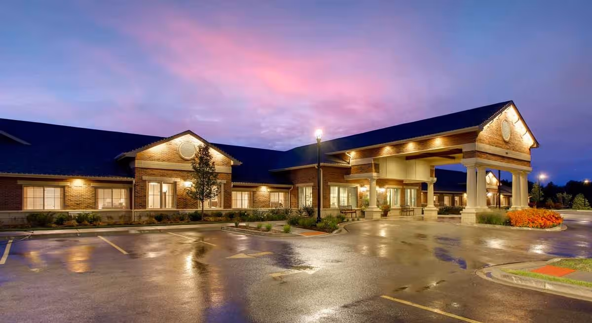 Exterior front view of a single-story brick memory care facility at dusk with a covered drop-off entrance and a wet reflective parking lot.
