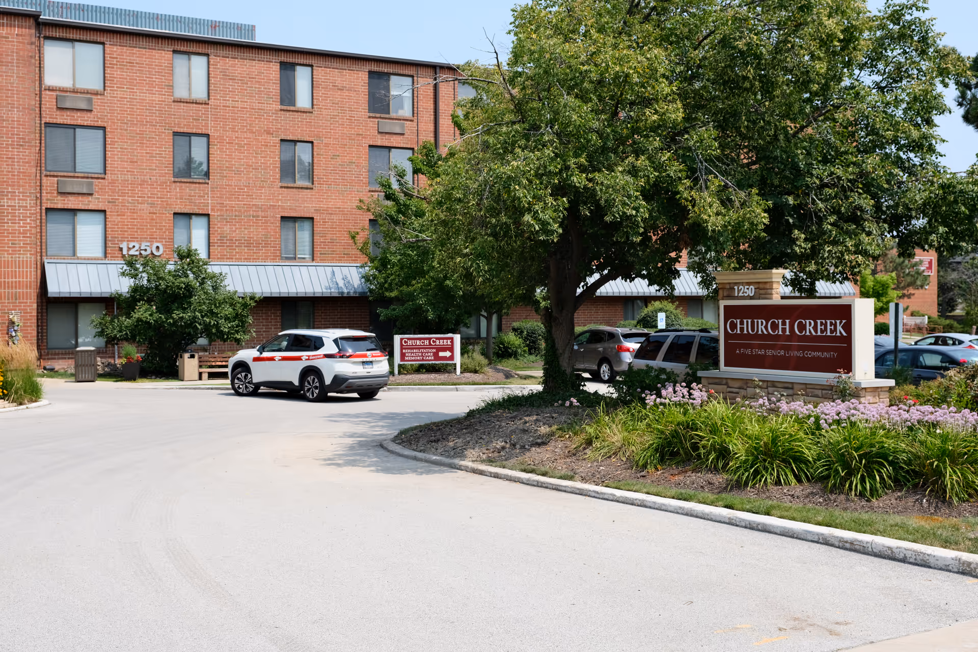 Exterior view of Church Creek senior living community building with a parking area, cars, trees, and a sign displaying the facility name and address 1250.