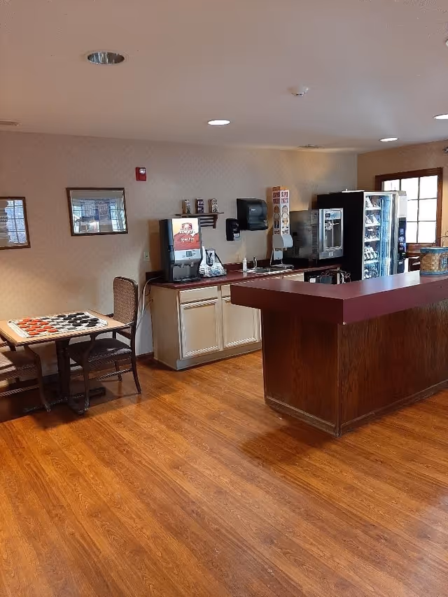 Interior view of a common area with a wooden floor, a small table with two chairs and a checkers board, a counter with a maroon top, beverage dispensers, a vending machine, and framed pictures on the wall.