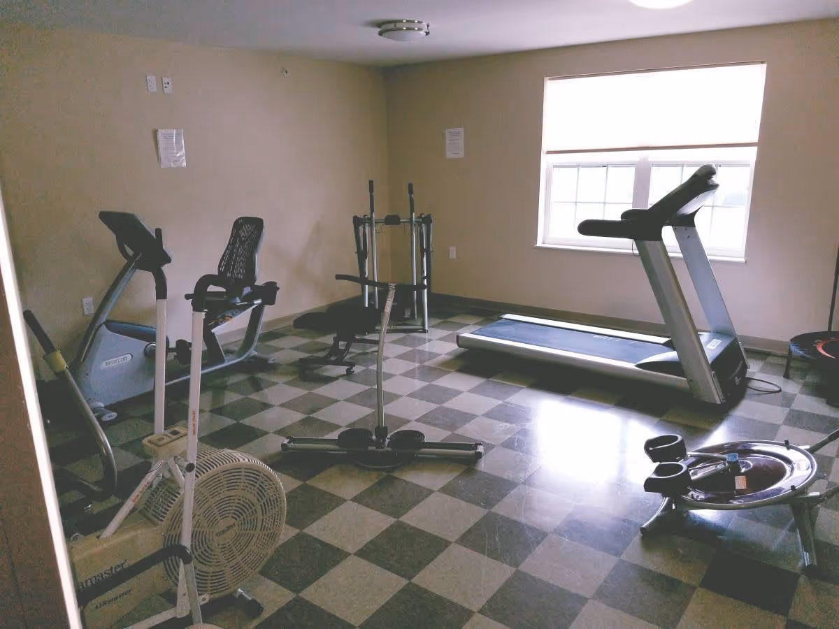 A small fitness room with exercise equipment including a treadmill, stationary bike, rowing machine, weight bench, and other workout machines. The room has checkered flooring and a window letting in natural light.