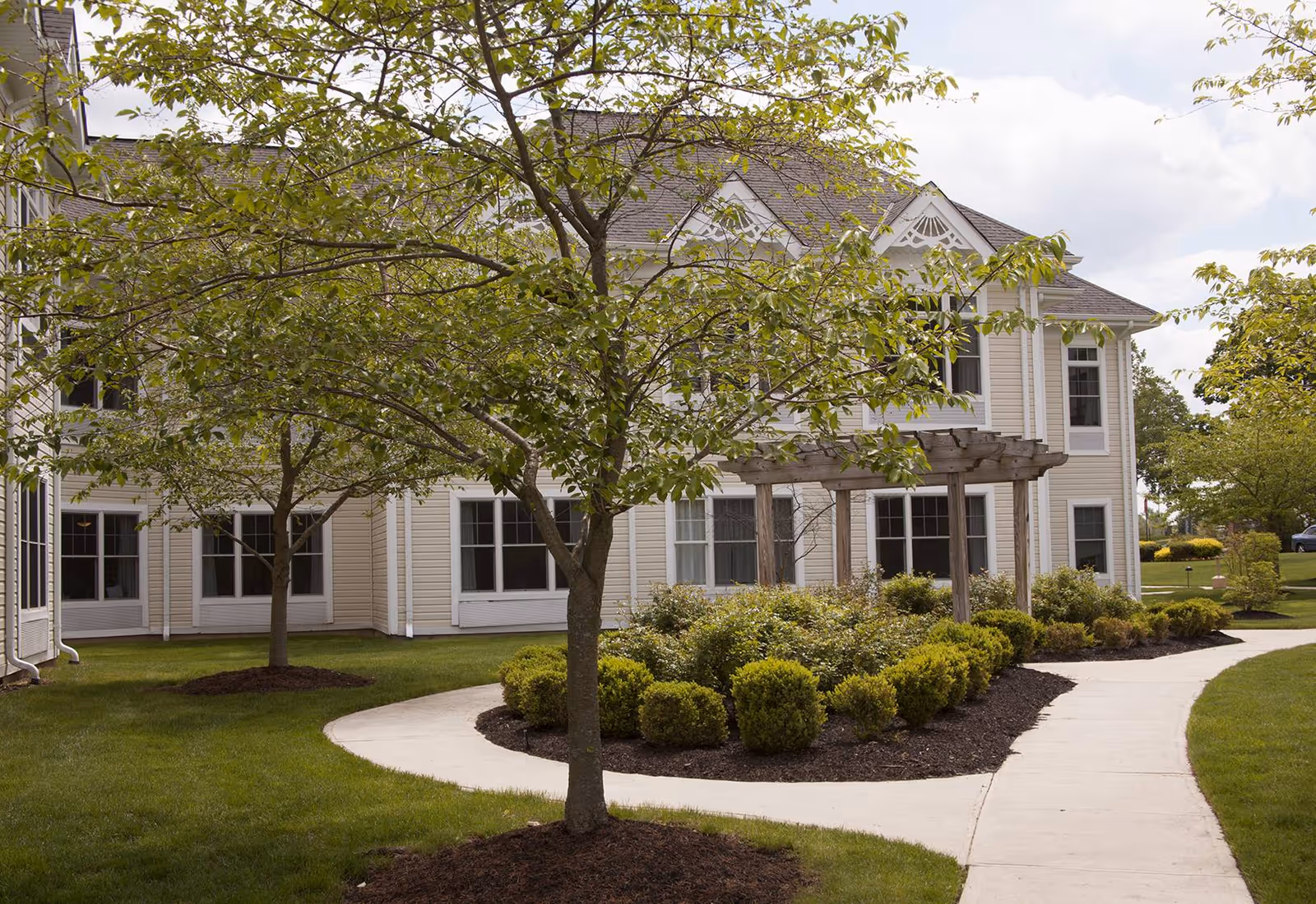 A landscaped outdoor area at Sunrise of Madison featuring a curved concrete walkway, green grass, neatly trimmed bushes, and several trees with green leaves. The background shows a light-colored two-story building with multiple windows and a wooden pergola structure.