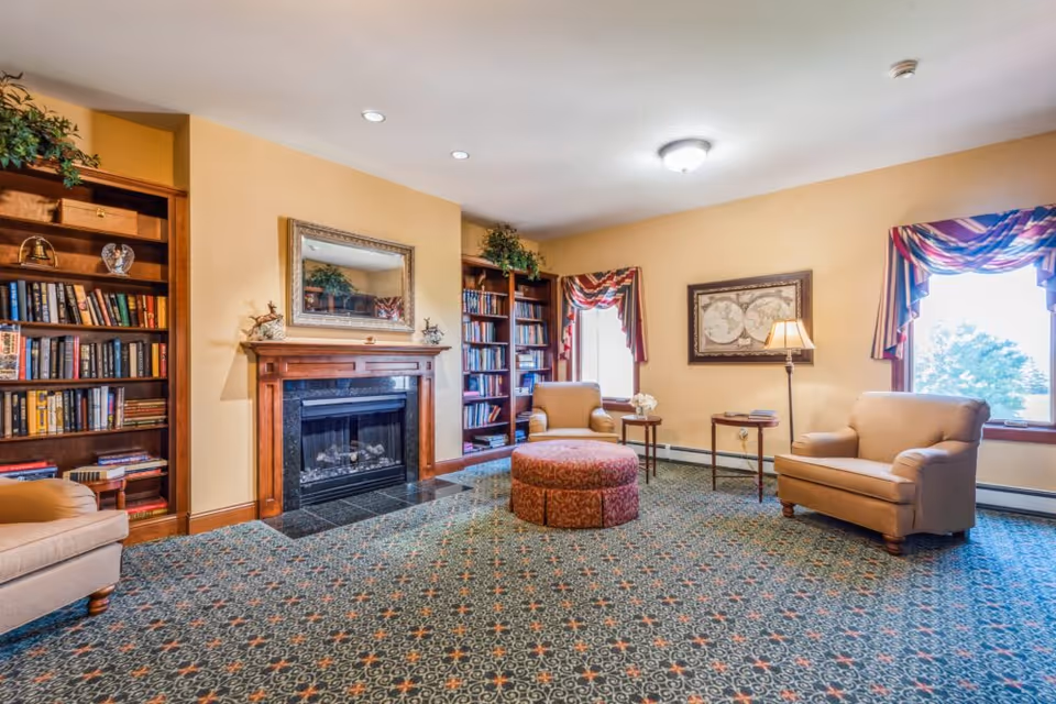 A cozy senior living room with beige walls and patterned carpet. The room features a wooden fireplace with a mirror above it, flanked by two tall bookshelves filled with books and decorative items. There are three beige armchairs arranged around a round, patterned ottoman. Two windows with colorful valances allow natural light to enter the room. A floor lamp and framed artwork decorate the space.