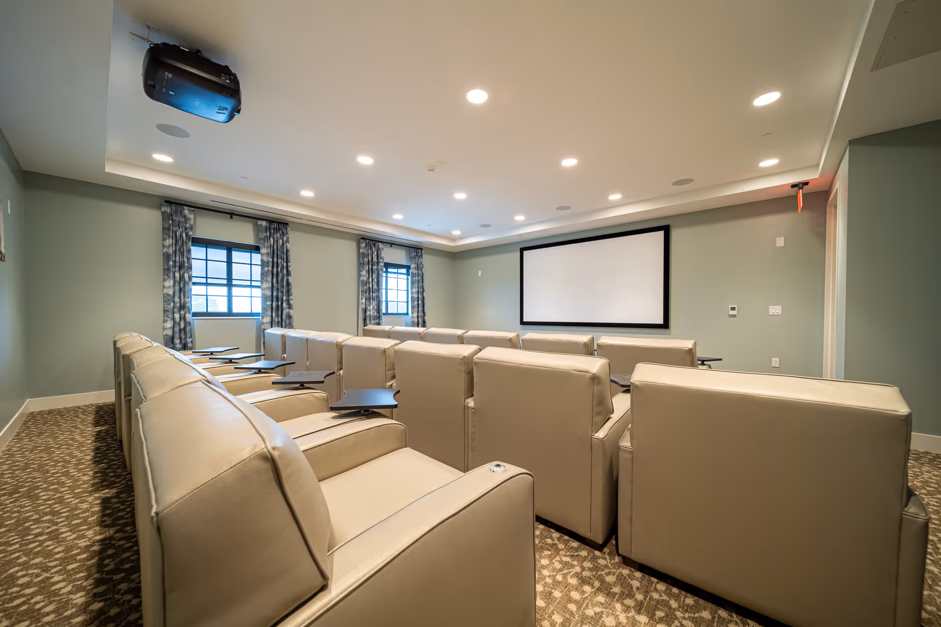 A modern movie theater room with beige leather recliner seats arranged in rows facing a large blank projection screen. The room has light green walls, patterned carpet, two windows with blue patterned curtains, and ceiling lights. A projector is mounted on the ceiling.