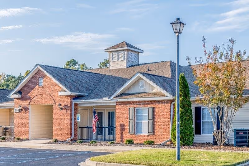 Exterior view of a single-story senior living facility building with brick and siding walls, a small covered porch with an American flag, a street lamp, and landscaped greenery including trees and bushes under a clear sky.