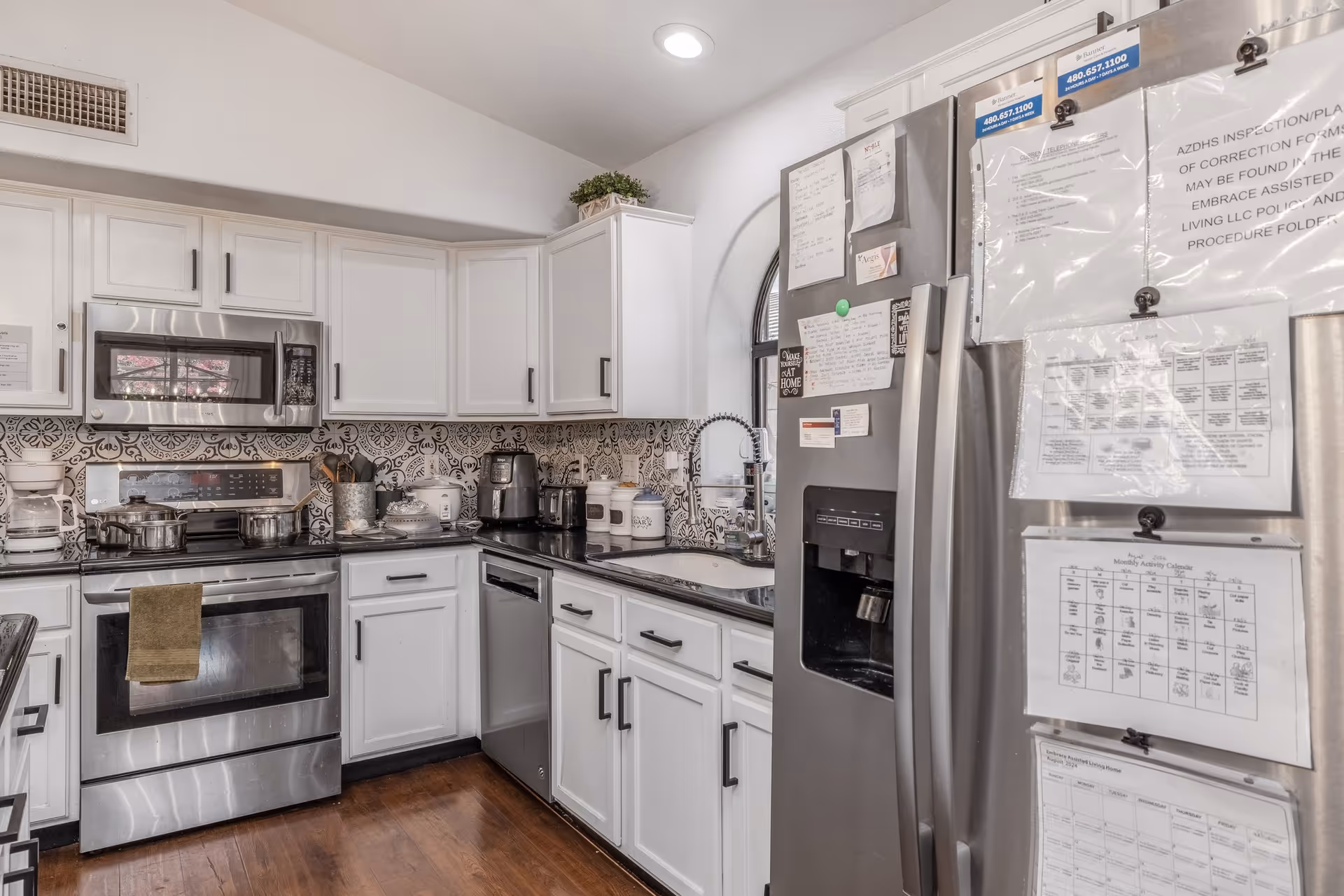A modern kitchen with white cabinets, stainless steel appliances including a microwave, oven, dishwasher, and refrigerator. The backsplash features a decorative black and white tile pattern. Various kitchen items such as pots, a coffee maker, and containers are on the countertops. Papers and notes are attached to the refrigerator door. The floor is wooden and the kitchen is well-lit with a ceiling light.