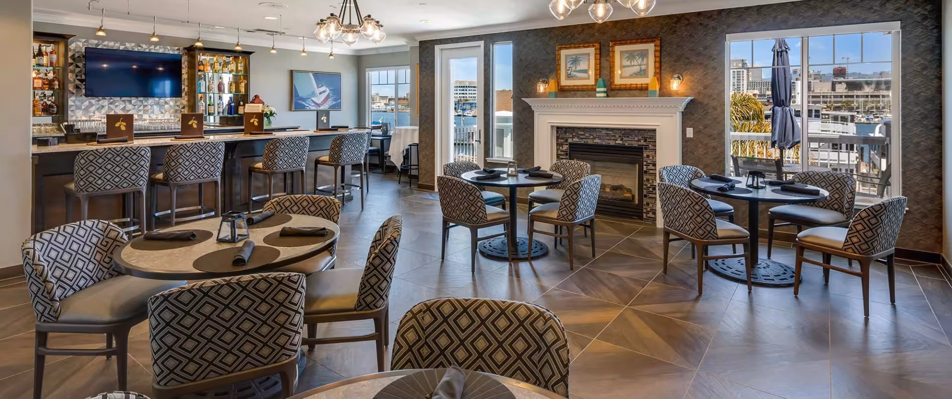 A stylish dining area with round tables and patterned chairs, a bar with stools and bottles of liquor, a fireplace with framed artwork above it, and large windows showing a waterfront view with boats and buildings outside.