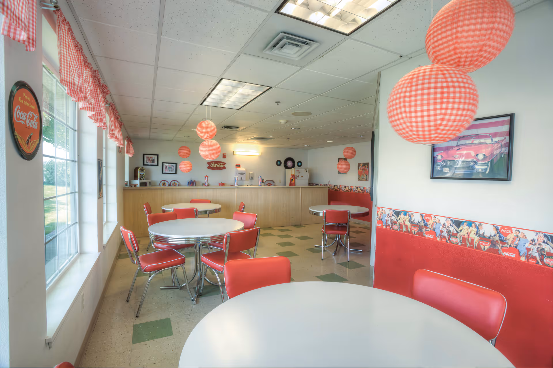 A retro-themed dining area with round white tables and red chairs. The room features large windows with red and white checkered valances, red and white checkered hanging paper lanterns, and vintage Coca-Cola decorations including wall art and a border along the wall. The floor has a checkered pattern with green and beige tiles, and there is a counter area at the back with various items on it.
