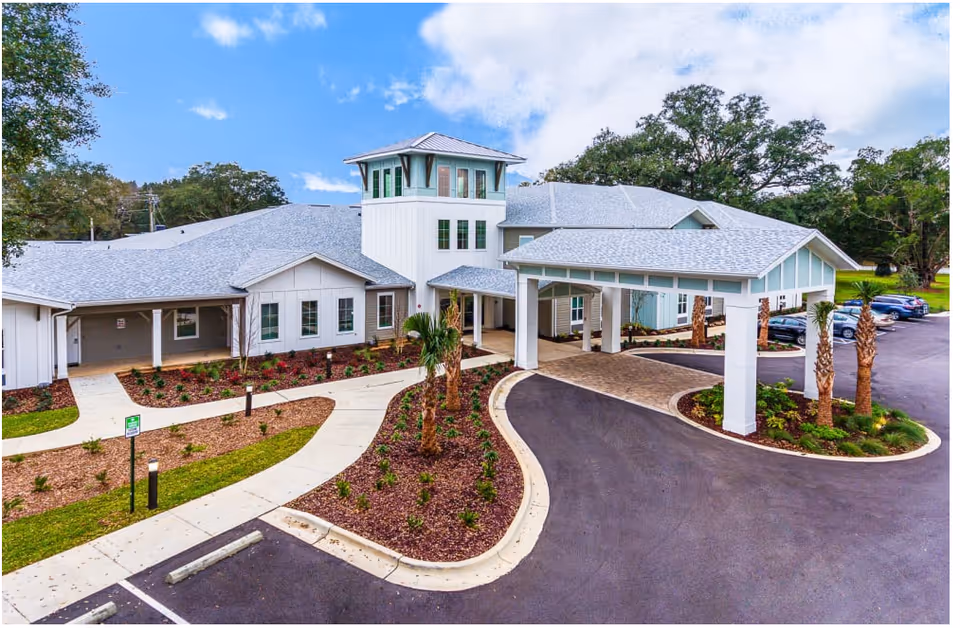 Front exterior view of a senior living building with a covered porte-cochere, landscaped entryway, and adjacent parking lot.