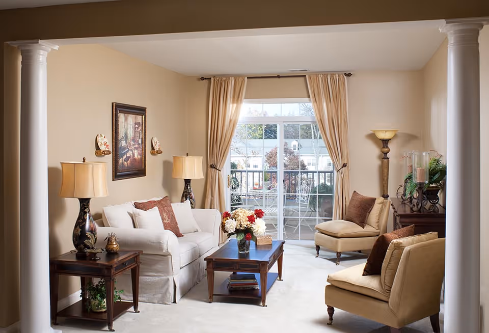 A cozy living room with beige walls and carpet, featuring a white sofa with decorative pillows, two beige armchairs with brown cushions, a wooden coffee table with a flower vase, two matching table lamps on side tables, a framed painting on the wall, and large sliding glass doors with beige curtains leading to an outdoor patio with white metal furniture.