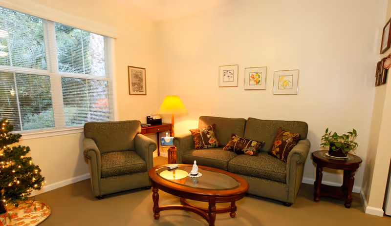 A cozy living room with a green upholstered armchair and matching sofa adorned with floral patterned pillows. A wooden coffee table with a glass top sits in front of the sofa, holding a small decorative item. To the right, there is a round wooden side table with a potted plant. A floor lamp with a yellow shade is placed near the corner, next to a small wooden desk. Three framed floral artworks hang on the wall above the sofa. A window with blinds shows greenery outside, and a small decorated Christmas tree is partially visible on the left side.