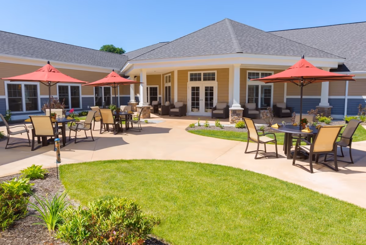 Outdoor patio area at Cornerstone at Milford Assisted Living & Compass Memory Support featuring several tables with red umbrellas and chairs on a paved surface, surrounded by green grass and landscaped plants, with a building in the background under a clear blue sky.