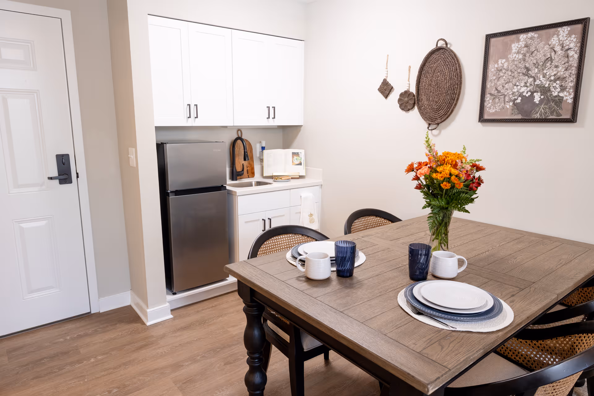 A small dining area with a wooden table set for two, featuring white plates, blue glasses, and white mugs. A vase with orange and red flowers is placed in the center of the table. Behind the table is a kitchenette with white cabinets, a small stainless steel refrigerator, a sink, and a cookbook on the counter. The wall is decorated with a framed floral painting and woven wall hangings. The floor is wood, and a white door with a black handle is visible to the left.