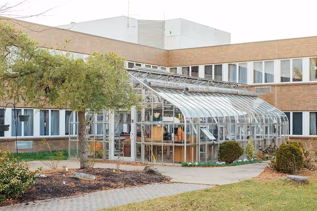 Exterior view of a senior living facility showing a glass greenhouse structure attached to a brick building. The surrounding area includes a paved walkway, grass, bushes, and a tree.