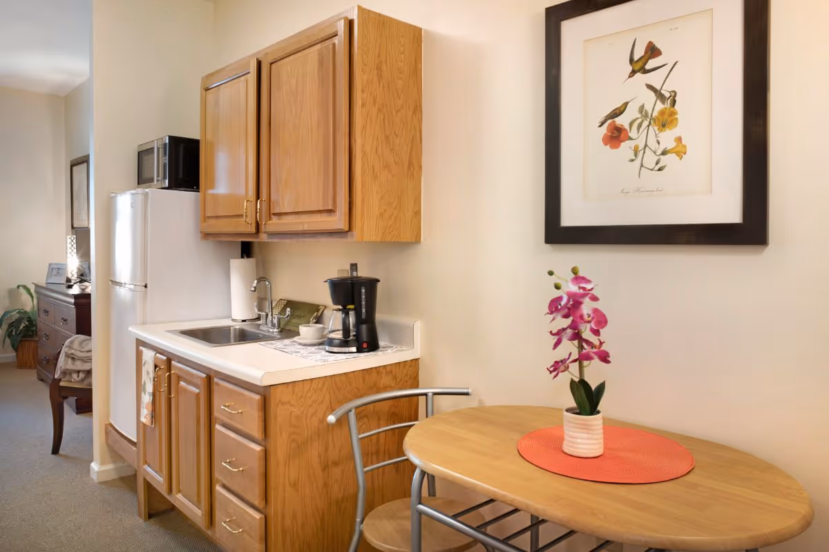 Small kitchenette with wooden cabinets, a sink, coffee maker, mini-fridge and a small table topped by a pink orchid and framed bird artwork.