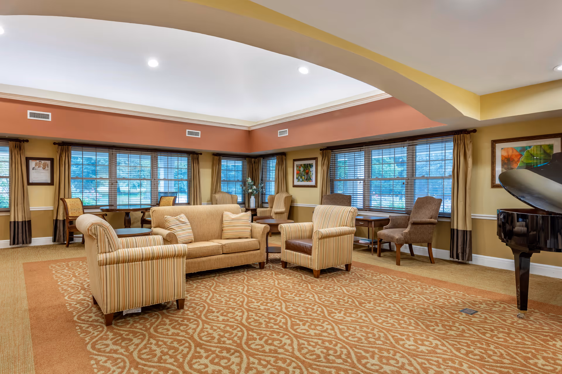 A spacious and well-lit living room area with a beige sofa, two striped armchairs, and additional seating near large windows with blinds. The room features a patterned carpet, warm yellow and orange walls, framed artwork, and a grand piano on the right side.