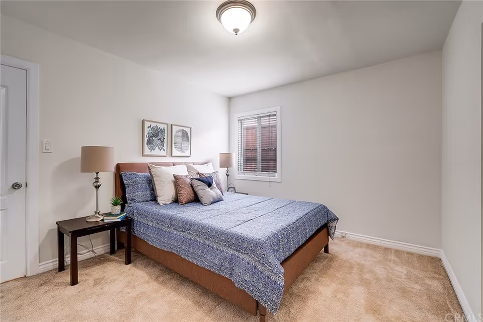 A bedroom with a bed covered in a blue patterned bedspread and multiple pillows. There are two framed artworks on the wall above the bed, a window with blinds on the right wall, and two bedside tables with lamps on either side of the bed. The room has beige carpet and white walls.