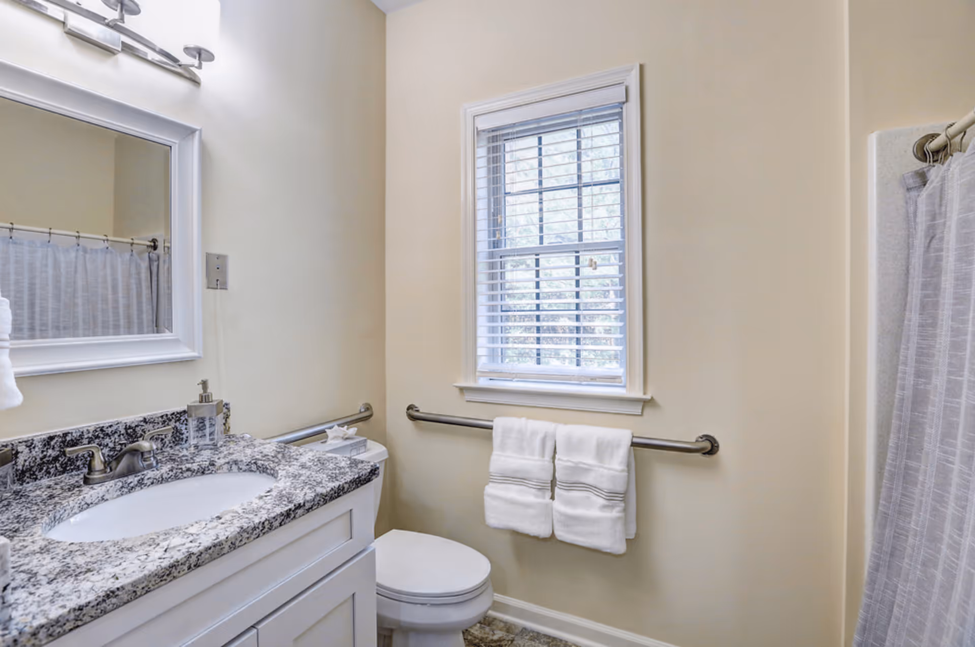 A clean bathroom with a granite countertop sink, a large mirror above the sink, a toilet, a window with blinds, and a towel rack holding two white towels. There is a shower with a curtain on the right side.