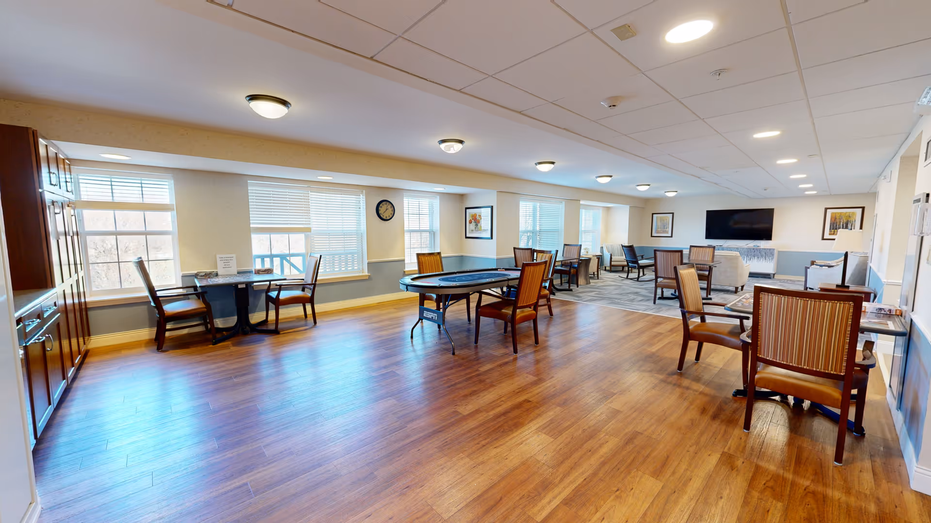 Bright spacious senior living common room with wood floors, multiple tables and chairs, and a wall-mounted TV.