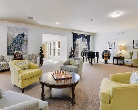 A spacious senior living common area with comfortable armchairs arranged around a round wooden coffee table with a chess set on it. The room features a grand piano, decorative plants, framed artwork, and large windows with dark blue curtains allowing natural light to fill the space.