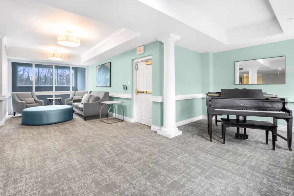 A bright and spacious senior living common area with light green walls, gray carpet, and white trim. The room features a gray sofa, two gray armchairs, a large blue oval ottoman, a small side table, a black upright piano with a bench, and a large mirror above the piano. There are three ceiling lights and large windows letting in natural light.