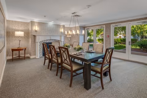 A well-lit dining room with a rectangular wooden table surrounded by eight cushioned chairs. The room features a fireplace with a white mantel, wall sconces, a floor lamp on a small side table, and large glass doors that open to a green outdoor area.