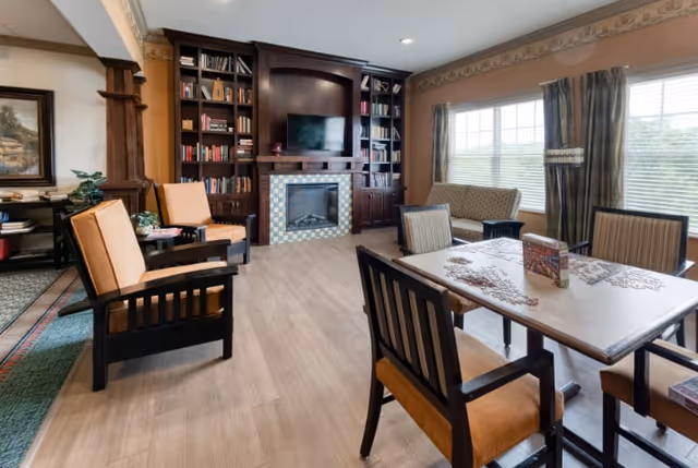 A cozy living room area in Cedarhurst of Columbia featuring a fireplace with a tiled surround and built-in dark wood bookshelves filled with books. There are comfortable chairs with orange cushions, a table with a puzzle in progress, and large windows with curtains letting in natural light.