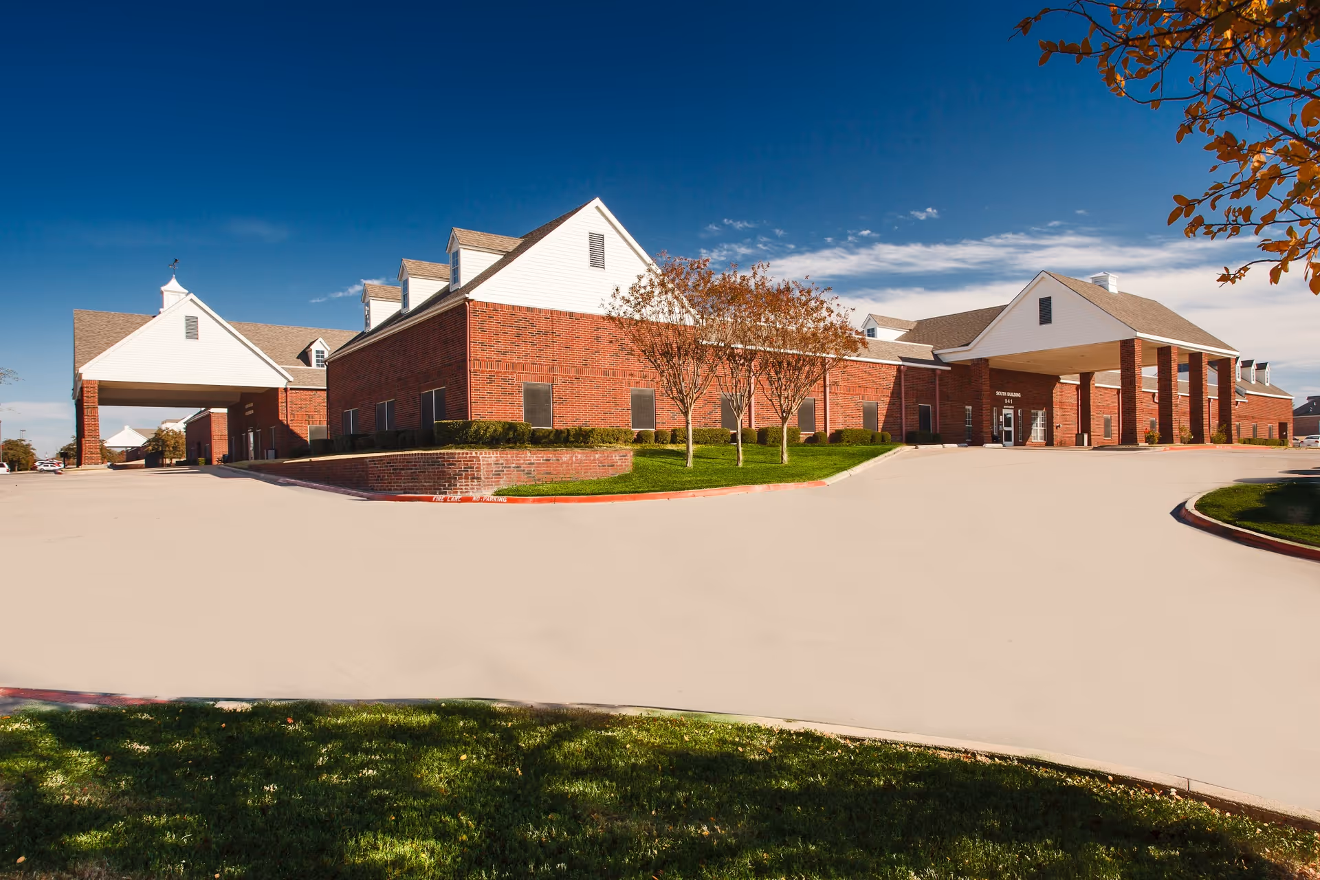 Exterior view of Williamsburg Village Healthcare Campus showing a large brick building with white gabled roofs, a covered entrance supported by columns, a driveway, and some trees with autumn foliage under a clear blue sky.