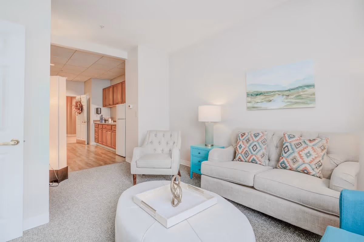 A bright and cozy living room area with a beige sofa adorned with patterned cushions, a matching armchair, a turquoise side table with a lamp, and a round ottoman with a decorative tray. In the background, there is a small kitchen area with wooden cabinets, a refrigerator, and a hallway with a door decorated with a wreath.